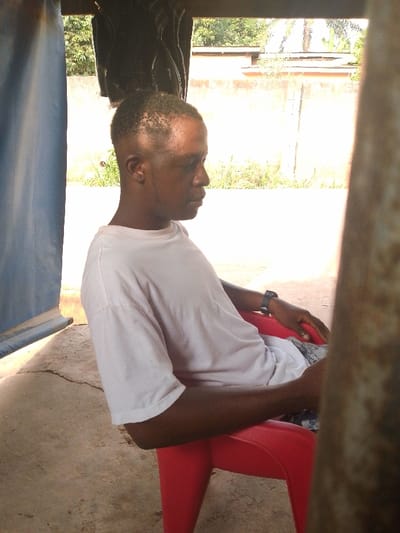 A man with short hair sits on a red plastic chair. He wears a white t-shirt and a black watch on his left wrist. The man looks towards the right. A concrete floor is beneath the chair. In the background is a wall, trees, and part of a building. A blue cloth hangs in the top left of the frame. The right side of the image is slightly blurred, possibly by an object close to the camera.