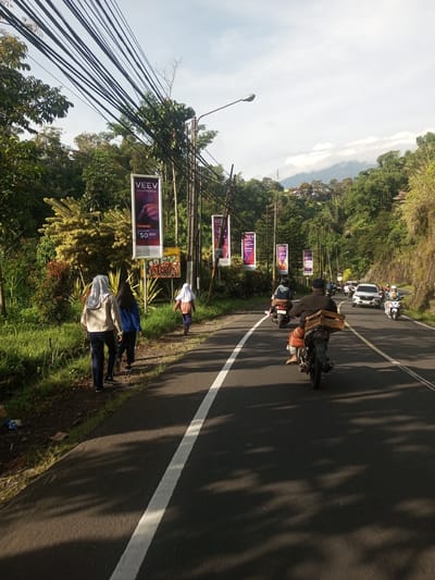 The image depicts a road scene in what appears to be a rural or semi-rural area. The road has a solid white line indicating the lane division. There are several people walking on the left side of the road, along the shoulder. They are walking away from the camera. On the road, several motorbikes and a white car are traveling in the same direction. A man on a motorbike is carrying a box. Along the road, there are several advertising banners with the word "VEEV" on them. Lush greenery and trees are present on both sides of the road. In the distance, hills and possibly a mountain are visible under a partly cloudy sky. There are many electric lines above the road. There is no indication of Phou Khoun, Laos.