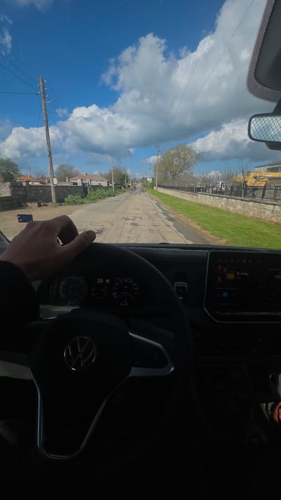 A person is driving a Volkswagen vehicle on a cracked asphalt road in Balgarevo, Bulgaria. The view through the windshield shows a street lined with houses and telephone poles under a blue, cloudy sky. The car's dashboard and steering wheel are visible, and a hand is seen holding the steering wheel. The navigation screen displays "Te Kelas" by Coke Studio. The environment appears rural or suburban.