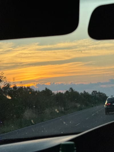The image captures a sunset viewed from inside a vehicle. The sky displays vibrant hues of orange and yellow, with wispy clouds stretching across. Below the sky, a line of dark trees forms the horizon. A paved road runs diagonally through the scene, with white lane markings visible. A dark-colored car is traveling on the road in the distance. The foreground shows parts of the vehicle's interior, including dark-colored elements that obscure the bottom and top edges of the frame. The overall setting appears to be a rural or suburban area, possibly near Parvomay, Bulgaria, given the context provided.