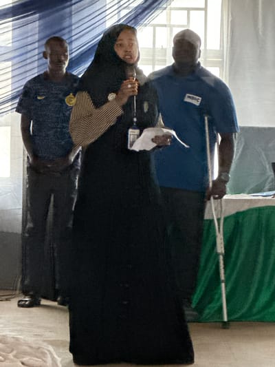 The image shows three individuals standing indoors, likely at an event or gathering in Unguwan Dosa, Nigeria. The setting appears to be a room with natural light coming from windows partially covered by blue striped curtains or fabric on the left side.

In the foreground, a woman wearing a black hijab and a striped top is centrally positioned. She holds what appears to be a microphone or a similar handheld device in her right hand, suggesting she might be speaking or presenting. A ring is visible on her finger. To her right stands a man with a bald head, dressed in a blue polo shirt that features a white rectangular logo on the chest, possibly reading "Victor." He is holding a long, thin white pole or stick in his right hand and appears to be holding a small white document or paper in his left hand, near the woman. To the woman's left, another man with a bald head is visible, wearing a dark blue or black shirt with a round gold badge or logo on the chest.

The background includes a large, dark-colored structure, possibly a display stand or backdrop. Further to the right, a significant portion of green fabric is visible, draped and folded, strongly suggesting it is part of a national flag, such as the Nigerian flag, given the context. The overall atmosphere appears semi-formal, indicative of a public event or presentation. The image has a slightly blurry quality with some visual noise.