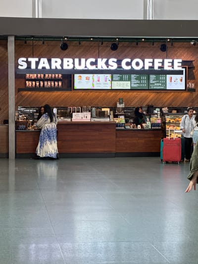In Tambon Mai Khao, Thailand, a Starbucks coffee shop is visible within an airport terminal. The store features a large illuminated sign that reads "STARBUCKS COFFEE". Customers are interacting at the counter, with some purchasing items. A variety of food and drink items are displayed behind the counter and in a refrigerated case. Two individuals are visible interacting with a barista, while another person is waiting in line. Luggage, including a red suitcase, is present near the counter. The floor is tiled and reflective. The environment appears clean and well-lit.
