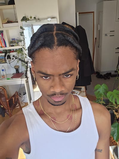 A person is posing indoors. The individual has dark skin, short, braided hair, and gold hoop earrings. They are wearing a white tank top and gold necklaces. There are red marks on their neck and chest. The background shows indoor items such as a potted plant, furniture, shelves with various objects, and a window. There is no evidence that this image was taken in Pantin, France.