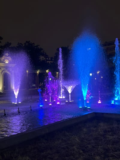 This is a nighttime, eye-level, medium shot of a public fountain in Bilbao, Spain. The fountain is illuminated by vibrant purple and blue lights, casting a colorful glow on the water and surrounding area. Several water jets shoot upwards, creating dynamic shapes and patterns against the dark sky. In the background, trees and building facades are visible, dimly lit by streetlights or the fountain's illumination. A few indistinct figures are present in the mid-ground, suggesting the fountain is a public gathering spot. The foreground shows a dark, grassy area bordered by a concrete edge.