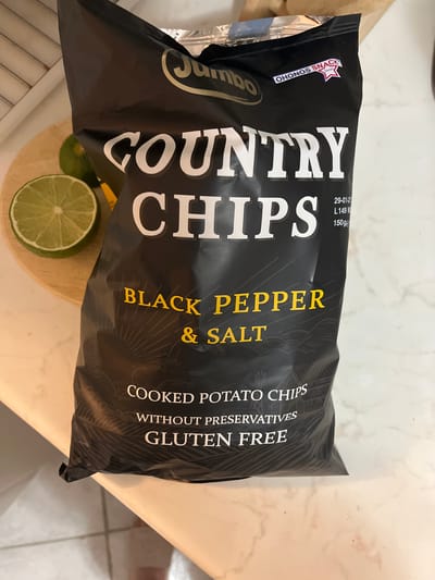 A bag of Jumbo Country Chips "Black Pepper & Salt" cooked potato chips is lying on a marble countertop. The bag is mostly black with white and yellow text. There is a lime cut in half to the left of the bag. The background appears to be a kitchen environment. There is no visible indication that this image is located in Gerakiní, Greece.