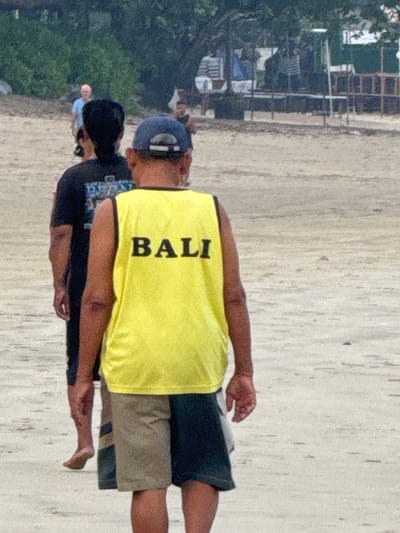 On a sandy beach in Jimbaran, Indonesia, a man in a yellow "BALI" tank top and dark shorts stands barefoot. Another person in a dark shirt stands nearby, also barefoot. In the background, trees and buildings are visible.