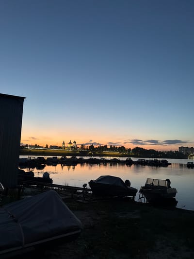 A tranquil sunset scene unfolds over a body of water, likely the Volga River in Syzran, Russia, as indicated by the presence of a cityscape featuring church domes and a power plant stack. The sky transitions from a deep blue overhead to warm hues of orange and pink near the horizon, painted with scattered clouds. A line of boats, some covered and others partially visible, are docked along the shore. In the foreground, a large structure, possibly a boathouse or industrial building, dominates the left side of the frame, its dark facade contrasting with the illuminated sky. The water reflects the colors of the sunset and the lights from the distant shore, creating a shimmering effect. The overall atmosphere is one of quietude and natural beauty as day transitions into night.