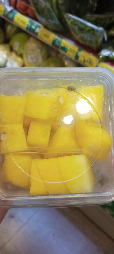 The image captures a close-up view of a clear plastic container filled with bright yellow, pre-cut fruit chunks, likely pineapple. Condensation is visible on the inner surface of both the container and its transparent lid.
A prominent detail is a small, dark-colored insect, resembling a beetle or a bug, resting on the inside of the container's lid, near the top right. Several small white particles are also visible among the fruit pieces. The container is held by a person, with a part of their hand visible at the bottom.
The background, largely out of focus, suggests a supermarket or grocery store environment. It shows various other produce items, some individually wrapped in plastic bags, arranged on shelves. Green and yellow price or information strips are visible on these shelves.
Visible text on the background strips includes numerical values such as "11.900", "7.000", and "7.300". Given the location context of Cipanas, Indonesia, these numbers most likely represent prices in Indonesian Rupiah. The scene indicates a shopping activity where a customer has observed a quality control issue with a packaged food item. The time of day cannot be determined, but the setting is indoors.