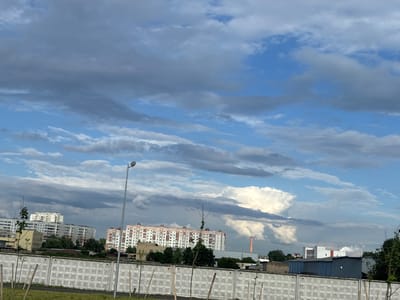 The image shows a cloudy sky over an urban landscape, likely within the Сеніцкі сельскі Савет, Belarus. In the foreground, there is a white concrete wall with a repeating patterned texture, reinforced with thin wooden poles planted in the ground in front of it. Beyond the wall, there are low-lying green bushes and some young trees.
Further back, several apartment buildings are visible. The one on the left is a multi-story building with a light-colored facade and numerous windows. To its right, a larger, pink and white apartment building stands out with a row of windows and decorative elements along its roofline. In the background, industrial buildings and a tall, orange chimney are present. The sky is filled with a mix of dark gray and lighter, wispy clouds, with patches of blue sky visible. A street lamp on a tall metal pole is situated to the left of the frame. The overall impression is of an ordinary day in a residential and industrial area.