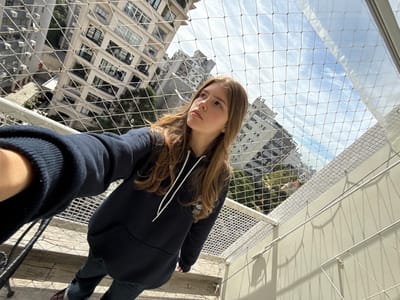 An elevated, eye-level selfie-style photograph depicts a young female standing on a high-rise residential balcony in Porto Alegre, Brazil. The balcony is secured by a light-colored, fine-mesh safety net with a grid pattern, prominently visible throughout the foreground and overhead.

The subject is a light-skinned female with long, wavy brown hair, positioned centrally but slightly angled to the right. She is looking upwards and to the viewer's left, away from the camera. She wears a dark blue hooded sweatshirt with white drawstrings and dark-colored pants. Her left arm, extended into the frame from the lower left, holds the camera for the self-portrait.

The balcony floor is light-colored and appears tiled. A dark, thin metal support structure, possibly part of furniture, is partially visible on the far left. The background reveals a dense urban environment with multiple multi-story, light-colored buildings displaying numerous windows, consistent with residential or mixed-use structures. Green foliage from trees is visible between these buildings. The sky above is partly cloudy, showing patches of blue sky and white clouds. Natural daylight illuminates the scene, casting distinct grid-like shadows from the safety net onto the adjacent building wall on the right side of the frame.