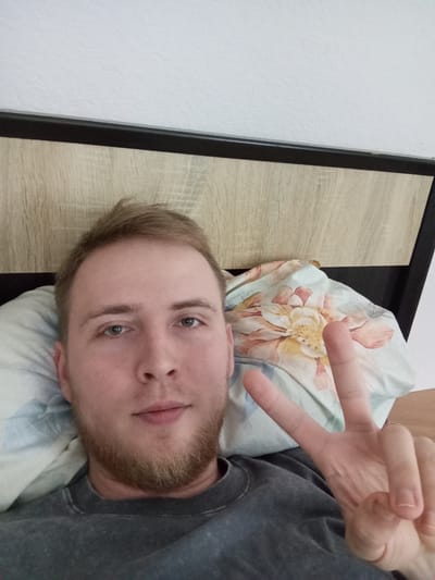 In a medium shot, a young adult male with light brown hair and a short beard lies in bed, facing the camera and raising his right hand in a peace sign. He is wearing a dark grey t-shirt and is positioned against a pillow with a floral pattern. Behind him, a wooden headboard and a white textured wall are visible. The image appears to have been taken indoors, possibly in a bedroom in Velyka Dymerka, Ukraine.