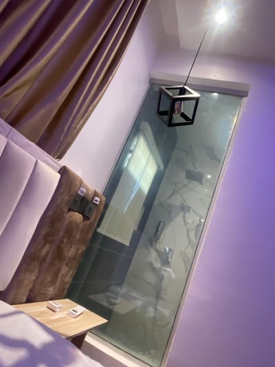 This image captures a modern interior room, likely an accommodation, situated in Galadimawa, Nigeria. The scene is observed from an angled perspective, revealing various room elements. On the left, a section of pleated, golden-brown curtains hangs above a light-colored, vertically upholstered headboard of a bed. A brown, possibly velvet, upholstered panel extends from the headboard, featuring two dark-colored wall-mounted electrical outlets or control panels with a small, partially legible white label above them. A white bedspread is visible in the lower left foreground.
A light-toned wooden nightstand is positioned next to the bed, holding a white cylindrical object and a small white remote control with colored buttons. The right side of the room is characterized by a smoothly painted purple-lavender wall. From the ceiling, a pendant light fixture descends, featuring a black cord and a geometric black cube-shaped frame that encloses a brightly illuminated bulb, emitting a distinct starburst effect. A large, transparent glass partition extends vertically, separating the main room from an adjacent space, which appears to be a bathroom or shower area. Through the glass, a white tiled wall with prominent dark, marble-like veining is visible, along with metallic shower fixtures and reflections of other internal bathroom elements. No individuals or specific interactions are present in the scene.