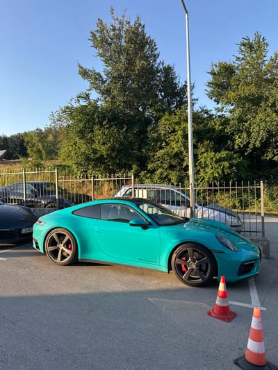 A turquoise Porsche 911 Carrera S is parked on an asphalt surface in the city of Kerenite, Bulgaria. The car is positioned in a parking space marked by white lines, with traffic cones on either side. In the background, a metal fence encloses a grassy area with trees and a building. Other cars are visible behind the fence, including a black Porsche and a silver vehicle. A tall metal pole, possibly a lamppost, stands near the car. The scene is bathed in sunlight, casting shadows on the asphalt. The sky is clear and blue.