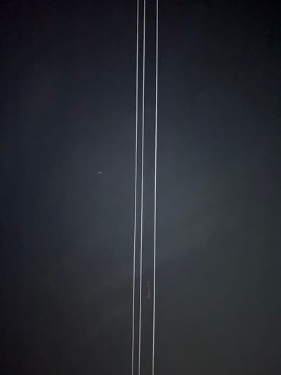 The image shows a dark, abstract background with three thin, vertical white lines positioned slightly to the left of the center. The lines appear to be of equal thickness and are spaced closely together. The background is mostly uniform in color, with some subtle variations in shading, suggesting a smooth, possibly artificial surface. There are a few small, faint white specks scattered across the background, which could be dust or imperfections. The overall impression is minimalist and somewhat enigmatic. The image does not contain any recognizable landmarks or elements that would indicate its location within Germasogeia Municipality, Cyprus, or any urban environment. It is a purely visual composition focusing on lines and a dark background.
