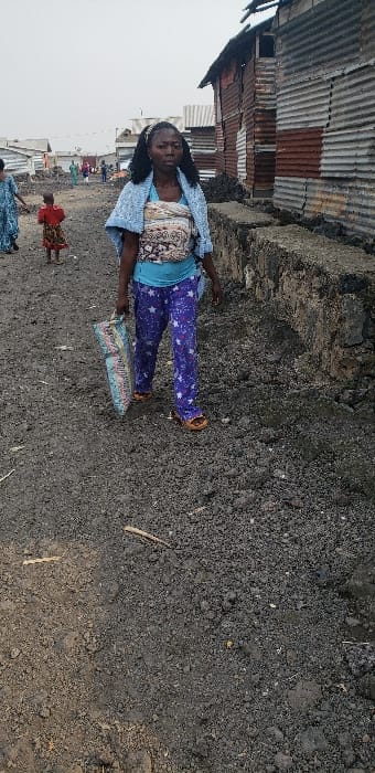 The image captures an outdoor scene in Buzove, Democratic Republic of the Congo, under an overcast sky. The primary subject is an adult female walking forward on an unpaved, dark, rocky path. She has dark skin and black braids, and is wearing a light blue sleeveless top, blue pants with white star patterns, and brown open-toed sandals. A light blue knitted shawl is draped over her shoulders, and a patterned fabric wrap is visible on her chest, securing a partially obscured infant or small child. In her left hand, she carries a rectangular, multi-colored bag. To the left and slightly behind her, another individual in a blue patterned dress is partially visible, and further back, a small child in a red top walks away. In the distant background, several other indistinct figures are present. To the right of the path, a series of rudimentary structures constructed from corrugated metal sheets, some exhibiting rust, line the area. A low, dark stone or concrete wall/curb runs parallel to these structures along the path.