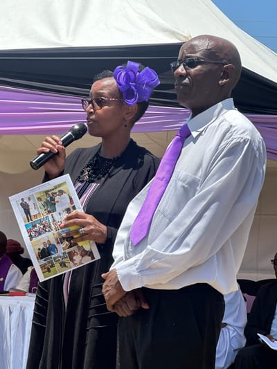 The image depicts a woman speaking into a microphone while holding a photo album and standing next to a man wearing a white shirt and a purple tie. The man stands with his hands clasped in front of him, his expression focused on the woman as she speaks. The woman is wearing a black and white patterned blouse, a black blazer, and a purple bow in her hair. The woman is wearing glasses. The photo album contains images of various people, possibly family or friends, with a title that reads "Deceased Wangari Group." The scene is taking place under a white tent with purple and white fabric decorations. It is likely a funeral or memorial service based on the photo album and the attire of the people present. The image is shot from a slightly low angle, focusing on the woman and the man. The background includes other people and a table with white cloth, suggesting the event is taking place in a public space.  Based on the provided coordinates, this scene is likely taking place in the town of **Embu, Kenya**, within a public space like a community hall or church grounds. The environment is bright and sunny.