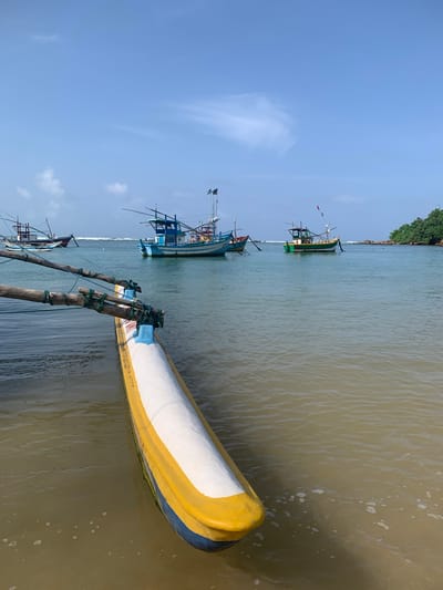 The image depicts a serene coastal scene in Mirissa, Sri Lanka, with a focus on traditional fishing boats. In the foreground, a brightly painted outrigger canoe, featuring yellow, blue, and white hues, rests partially submerged in the shallow, murky water near a sandy shore. Two wooden outriggers extend from the canoe, secured with rope.
In the middle ground, several other fishing boats are anchored in the calm, turquoise ocean. One prominent boat, painted blue with white accents and red trim, has two masts and several flags. Another boat, predominantly green with yellow and white details, also features masts and flags. Further in the background, another fishing boat with a red and white hull is visible. The water shows gentle ripples, suggesting a calm sea.
The horizon is marked by a lush, green headland dotted with trees, which extends into the sea on the right side of the frame. The sky above is a clear, bright blue with a few wispy white clouds. The overall atmosphere is tranquil and indicative of a typical day in a coastal fishing village.