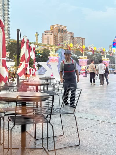 An outdoor scene in Nanshan District, China, features an employee wearing a gray cap and dark apron, standing next to stacked metal chairs on a tiled surface. Red and white striped umbrellas provide shade over outdoor tables. In the background, a hotel named Indigo and a Ferris wheel are visible along with several other buildings and people walking in the distance. Festive lights decorate the area.