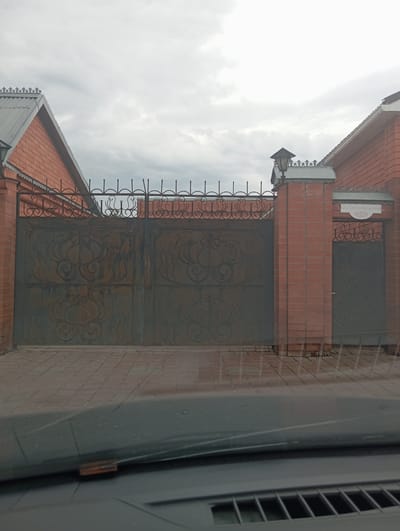 The image shows a brick house with a metal gate. The gate is made of dark metal with ornate designs. There is a light fixture on top of a brick pillar next to the gate. A sign with Russian writing is visible on the pillar. The ground in front of the gate is paved with bricks. The sky is overcast. The image appears to be taken from inside a vehicle, as the dashboard and windshield are visible in the foreground.