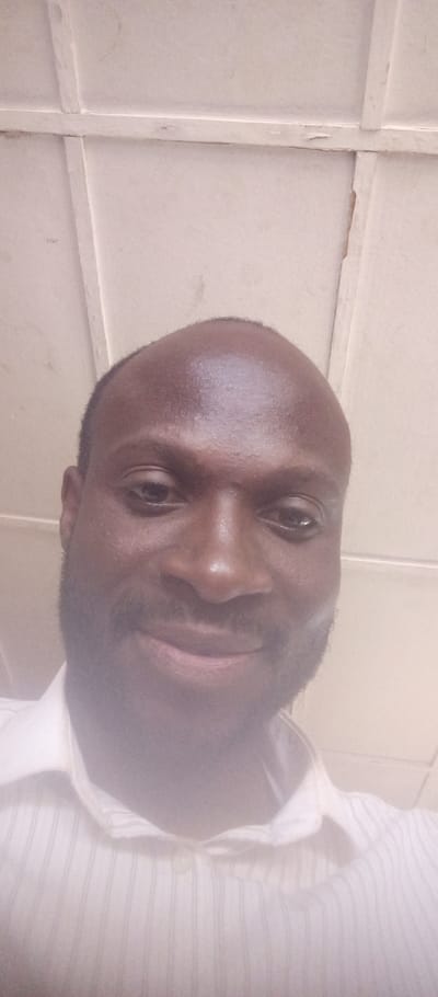 An upward-angled close-up portrait depicts a single adult male, positioned centrally. The male has dark skin, a short dark beard, and a receding hairline. He is wearing a light-colored, possibly white or off-white, collared shirt featuring faint vertical stripes. He looks directly into the camera with a slight smile. The background consists of an interior, light-colored (off-white) paneled ceiling, featuring visible rectangular sections and structural beams. Minor irregularities and discoloration are present on the ceiling surface. The scene is located in Barnawa, Nigeria.