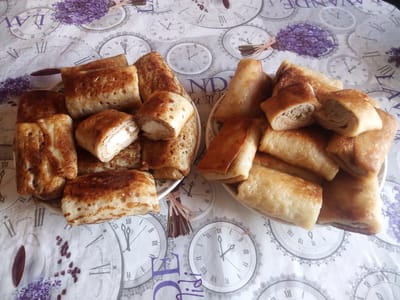 A top-down shot displays two plates of savory crepes, or blini, filled and rolled, arranged on a tablecloth adorned with vintage clock faces and sprigs of lavender. The crepes are golden brown and appear to be filled with a light-colored, creamy filling on the left, and a darker, meat-like filling on the right, visible in the cut sections of some crepes. The tablecloth features Roman numerals and decorative clock hands, suggesting a theme of time or antique aesthetics. The overall presentation is casual, possibly a home setting, with no individuals or discernible city landmarks from Usman, Russia, present in the frame.