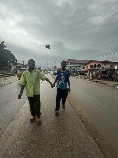 Two young boys are walking hand-in-hand on a wet, paved road in Epe, Nigeria. The sky is overcast and the weather appears to be gloomy, possibly after rain. The boys are dressed casually, with one wearing a blue t-shirt with a white design and dark pants, and the other in a lime green button-up shirt and dark pants. Both are wearing sandals.
In the background, there are buildings lining the street, which appear to be shops or businesses. Some of these buildings have awnings and visible wares. A yellow tricycle, common in this region, is visible on the road. Utility poles and wires are also present, indicating an urban or semi-urban environment. The road surface shows signs of wetness and some debris. The overall scene suggests an ordinary day in a bustling Nigerian town under a cloudy sky. No specific emotions are clearly discernible from the image, though the act of holding hands might suggest a bond or companionship between the boys. There is no visible text in the frame that is clear enough to be read.