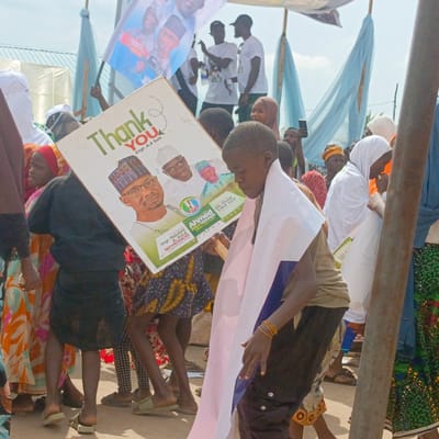 A crowd of people is gathered outdoors in what appears to be a political rally or demonstration in the city of Gudi, Nigeria. In the foreground, a child holds a large poster of Engr. A.A Sule, who is running for the office of Governor of Nasarawa State. The poster says, "Thank you, Engr. A.A. Sule," and includes photos of the candidate and his associates. The poster also mentions Senator Ahiyu Wada and Alh. Khalid Yusuf Sule.
The child also holds a cloth or flag. Behind the child, other people are visible, some wearing traditional African clothing. Further in the background, there are individuals wearing white shirts and standing beneath blue fabric and a large banner.
