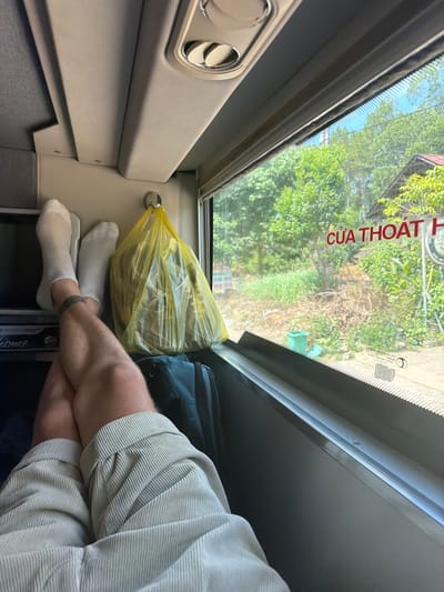 In Đại Ninh, Vietnam, a person's legs, clad in beige corduroy shorts and white socks, are crossed at the knees, resting on a dark blue suitcase. The legs are positioned in front of a window, looking out at a lush, green landscape with trees and a glimpse of a building with a red roof. A yellow plastic bag hangs from a hook on the wall next to the person. The interior appears to be a vehicle, possibly a bus, given the overhead lighting and window design. Red lettering on the window reads "CỬA THOÁT H", which translates to "EXIT DOOR".