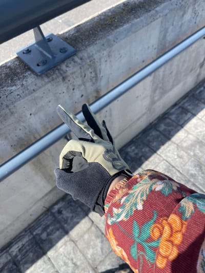 The image displays a close-up, downward-angled view of a person's right arm and hand in Altafulla, Spain. The hand, encased in a two-toned glove (black palm and inner fingers, light olive green/khaki back and outer fingers with a black Fox Racing logo), performs an "OK" gesture with the thumb and index finger forming a circle and the remaining three fingers extended upwards. The visible sleeve of the person's garment is a red/maroon corduroy fabric patterned with green leaves and orange/yellow fruits. In the background, a concrete structure serves as a barrier or railing. A dark grey metal railing is secured to the top of this concrete with a grey metal bracket and four bolts. Below the hand, a thinner, light grey horizontal metal bar is attached to the lower section of the concrete wall. The ground beyond the concrete structure is paved with light-colored square or rectangular pavers. Strong sunlight illuminates the scene, casting distinct shadows from the hand and the concrete structures onto the pavement.