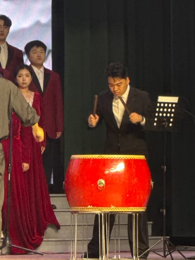 The image captures an indoor musical performance, likely on a stage, featuring a main drummer and other performers. In the central and upper part of the frame, a man dressed in a black suit and white shirt is actively playing a large, prominent red drum. The drum is adorned with decorative gold trim and rests on a metallic stand. The man holds a drumstick in each hand, demonstrating focused intensity on his performance.
Behind the drummer, to his left, is a black stand or equipment piece featuring a pattern of white circular lights or indicators. Further back, metallic scaffolding or stage rigging is partially visible.
In the lower foreground, three other individuals are present, presumably fellow performers. One man wears a maroon jacket over a white shirt, positioned next to a woman in a flowing red gown. Another person, dressed in a grey garment, is partially visible. These individuals have neutral expressions and appear to be part of the event, possibly observing or awaiting their cue.
The background is predominantly dark, suggesting a performance setting with controlled stage lighting. The attire of the performers and the style of the drum imply a formal, possibly cultural, musical presentation, consistent with the location context of Guta, China. No discernible text is visible within the image.