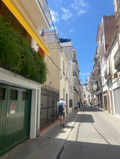 The image, rotated 90 degrees clockwise, captures a bright, sunny daytime scene down a narrow, pedestrian-friendly street lined with multi-story buildings in Sant Pol De Mar, Spain. The perspective looks down the street, with buildings towering on both sides, creating a canyon-like effect.
In the foreground on the left, a dense green bush sits above a bright yellow awning, partially obscuring the lower part of a white building. On the right, a prominent dark green wooden gate or garage door features decorative metal grilles and a red circular sign. This sign depicts a no-dumping symbol and text, which is clearer in a crop, reading "Sant Pol de Mar" and "No tiris la brossa" (Don't throw trash). Above the gate, a black metal fence with pointed spikes runs along the top of a low wall.
Several people are visible along the street. In the mid-ground on the right, a person in a white t-shirt, patterned shorts, and a hat is seen from behind, carrying a bag and wearing sandals, standing near the metal fence. Further down the street, other individuals in casual attire can be seen walking or standing, receding into the distance.
The street itself is paved with asphalt, flanked by patterned tiled pavements. Strong, distinct shadows are cast by the buildings and overhead wires onto the street and sidewalks, indicating high sun exposure, likely midday or early afternoon. A bright blue sky with sparse white clouds is visible between the buildings, with overhead wires crisscrossing the open space. Various building details are present, including balconies, windows, other awnings, and metal grilles. Signs for businesses are visible further down the street, including one that clearly reads "PIZZA" and another with vertical text that appears to say "BAR". The overall atmosphere is that of a bustling, vibrant Mediterranean town street.