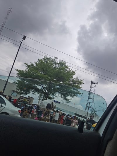 The view from inside a car in Benin City, Nigeria, shows a reflection in the windshield of a busy street. People stand along the sidewalk with goods displayed for sale; a white car is parked along the road. The sky is overcast. Overhead electrical wires run across the image. In the reflection, a communication tower can be seen. A large tree stands near the curb.