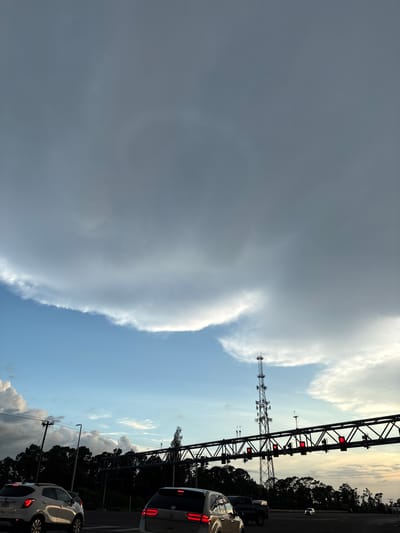 The image shows a view from a vehicle on a highway, likely approaching a toll plaza or an overhead gantry with traffic signals. The sky is filled with dramatic, heavy clouds, with a large, lighter-colored cloud formation dominating the upper portion of the frame. Below the clouds, a clear blue sky is visible, with the sun low on the horizon, suggesting either early morning or late afternoon.
In the mid-ground, a tall, lattice-style telecommunications tower stands to the left of center. A dark, industrial-looking overhead structure, possibly part of a highway overpass or a toll collection system, spans across the frame. This structure has multiple red lights illuminated, indicating traffic control signals. Trees line the roadside, appearing as a dark silhouette against the sky.
In the foreground, several cars are visible on the highway, suggesting moderate traffic. A white SUV is in the left lane, and the rear of a white SUV or crossover is in the right lane. The headlights and taillights of the vehicles are illuminated. The road surface is asphalt, and lane markings are visible.
The location is identified as Venice, United States. The overall scene conveys a sense of travel and the anticipation of reaching a destination, with the weather adding a dramatic element to the visual.