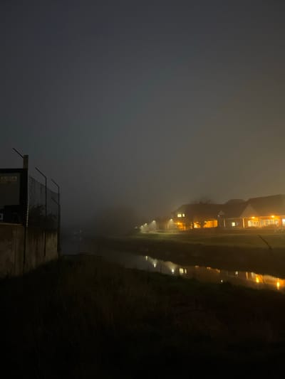 This forensic description details a foggy nighttime scene along a waterway in Lewes, United Kingdom. In the left foreground, a concrete wall and chain-link fence rise, partially obscuring a dark, indeterminate structure behind them. Yellow triangular warning signs with black pictograms are affixed to the fence structure. The immediate bank is covered with dark, indistinct vegetation. A calm body of water, likely a river or canal, extends horizontally across the midground, reflecting multiple points of artificial warm-toned light. On the opposite bank, a row of multi-story buildings, appearing to be residential or commercial, are visible. These structures exhibit illuminated windows and exterior lighting, casting warm light onto their facades and the surrounding ground. The entire scene is enveloped in dense fog, which diffuses light sources and significantly reduces visibility. The sky is dark grey, with a slight lightening effect near the horizon due to ambient light diffusion. No individuals or specific interactions are present in the frame.