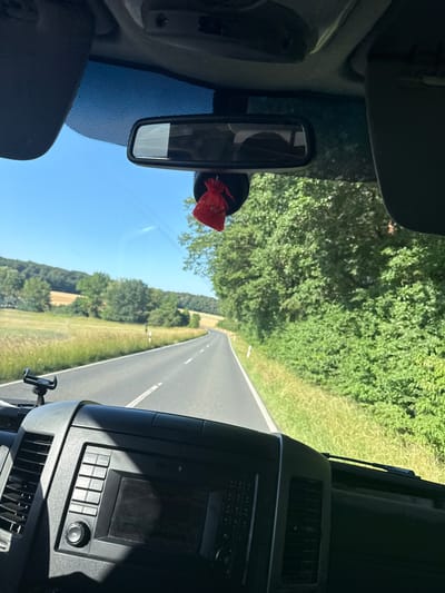 The image is taken from inside a vehicle, looking forward through the windshield. The view is oriented vertically, suggesting the photo was taken at an angle or the device was rotated. Outside, a paved road stretches into the distance, bordered by lush green trees and fields on either side. The sky is clear and blue, indicating sunny weather.
Inside the vehicle, the dashboard is visible on the right, with a radio or infotainment system and an air vent. A rear-view mirror hangs from the windshield, and a small red decorative item is attached to it. The interior suggests the vehicle is a truck or a similar large vehicle due to the dashboard's design.
The scene depicts a journey on a road through a rural area. The time of day appears to be daytime, given the bright sunlight. The location is identified as Oberwetz, Germany, which aligns with the European-style road and greenery. There are no people visible in the image. The overall mood is one of peaceful travel through a scenic, natural environment. There is no visible text in the image.