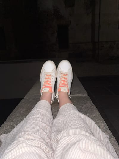 In Lagnasco, Italy, a person's legs are visible from the knees down, resting on a concrete ledge at night. They are wearing white sneakers with bright orange laces and loose, light-colored striped pants. The background is dark and out of focus, showing hints of a brick wall or building structure.