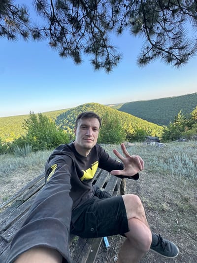 A man with short brown hair and a light complexion is sitting on a wooden bench outdoors, facing the camera and holding up two fingers in a peace sign with his right hand. He is wearing a dark long-sleeved shirt with yellow accents and black shorts. The bench is situated on a grassy slope overlooking a valley filled with lush green trees. The sky above is clear blue, and the branches of a tree hang into the top of the frame. The location is identified as the city of Pryvorottia Druhe, Ukraine.