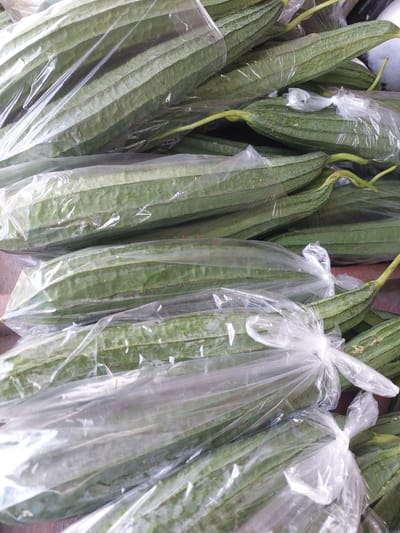 A pile of ridge gourds, also known as luffa or Chinese okra, are bundled together in clear plastic bags. The gourds are elongated and ribbed, with a deep green color. They appear to be fresh and ready for sale. The image was taken in a market in Parit Baru, Indonesia.