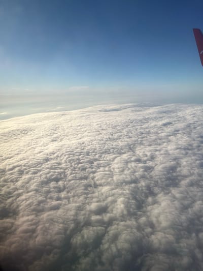 This is a high-angle photograph taken from an airplane window, showing a vast expanse of clouds below and a clear blue sky above. The image is oriented with the sky on the left and the clouds on the right. A small portion of a red and white airplane wing is visible in the upper left corner of the frame, indicating the perspective is from within the aircraft.

The clouds appear to be a dense, textured layer, possibly altocumulus or stratocumulus formations, with a somewhat undulating surface that creates shadows and highlights. The transition from the clear blue sky to the cloud layer is gradual, with a hazy area at the horizon. The light suggests it is daytime, likely during the morning or afternoon, given the brightness of the sky and the illumination of the clouds. The location is identified as Stolbishche, Russia, suggesting the flight is over this region. No people or text are visible in the image. The overall impression is one of aerial travel over a cloud-covered landscape.