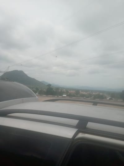 The image is a landscape scene taken from inside a vehicle, looking out towards a mountainous area and a village. The foreground is dominated by the roof of a white car, with black roof rails visible. The sky is overcast and cloudy, suggesting a potentially rainy or dull day.

In the mid-ground, a range of green mountains is visible under the cloudy sky. A large crane-like structure is partially visible on the left side of the image, near the base of the mountains. Below the mountains, a village or town is spread out, with buildings and trees dotting the landscape. A road or highway can be seen winding through the scene.

The overall impression is of a slightly hazy, diffused light, common on an overcast day. The setting appears to be rural or semi-rural, with natural elements like mountains and vegetation prominent, alongside human settlement. There is no visible text in the image. The time of day is difficult to ascertain precisely due to the overcast conditions, but it appears to be daytime. The mood conveyed is somewhat subdued, in line with the cloudy weather. The location is identified as Lankoviri, Nigeria.
