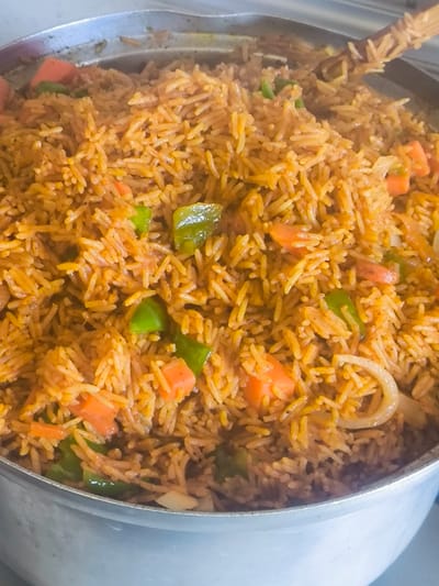 The image displays a close-up view of a large, metallic cooking pot filled with Jollof rice. The rice is a reddish-orange color, mixed with distinct pieces of diced orange carrots, chopped green bell peppers, and translucent white onion rings. Individual long-grain rice kernels are visible throughout the dish. A wooden utensil, possibly a spoon or spatula, is partially submerged in the rice on the upper right side of the pot. The pot's metallic rim is visible at the top and its curved side at the bottom of the frame. The background is a blurred, light-toned surface, indicative of an indoor setting, likely a kitchen. No individuals or active interactions are depicted. The specific location within Nicosia, Northern Cyprus / Cyprus, cannot be determined from the visual information provided.