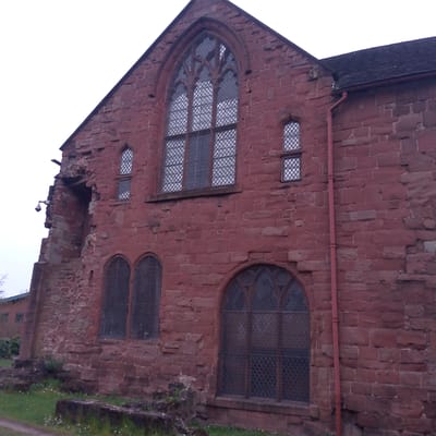 This image presents an angled view of a historic building constructed from red sandstone blocks, indicative of a structure like a church or monastic ruin. The facade features several distinct windows. A large, prominent Gothic-style arched window with intricate stone tracery and diamond-patterned leaded glass is centrally located in the upper section of the gable end. To its right, a pair of smaller arched windows with similar leaded glass are stacked vertically. On the lower level, there are additional arched windows, some of which appear to be partially boarded up or in a state of disrepair.
The left side of the building shows significant damage, with a large section of the wall missing, revealing an inner alcove or former opening. A modern security camera is mounted on the remnants of this damaged wall. A red downpipe runs vertically along the right side of the building, connecting to a dark-shingled roof that is partially visible against a bright, overcast sky.
The foreground consists of green grass interspersed with small white flowers and some low-lying stone foundations or debris. No people or specific activities are visible in the scene, which conveys a sense of stillness and historical presence. The lighting suggests an overcast daytime. There is no visible text within the image. The architectural style and materials are consistent with historical buildings found in the United Kingdom, specifically Coventry.