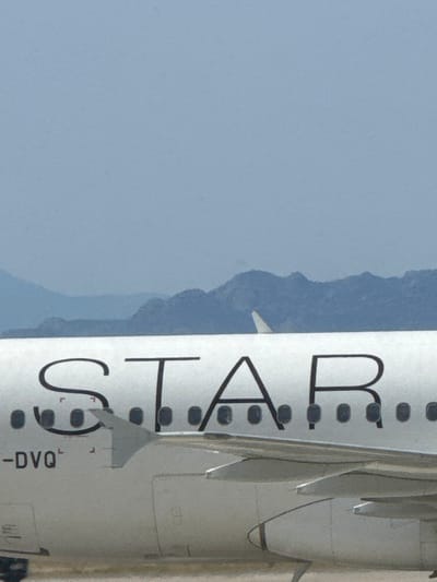 The image shows a close-up view of the side of a white airplane. The airplane is parked on a tarmac. The word "STAR" is printed on the side of the plane in black letters. The registration number "-DVQ" is visible on the fuselage of the plane. The airplane is located in the city of Paradisi, Greece, at the coordinates 36.403, 28.091. The background of the image is a hazy blue sky with mountains in the distance.