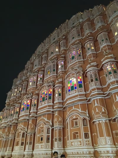 The image shows the Hawa Mahal in Jaipur, India, at night. The architectural facade features numerous small windows with intricate designs, some containing colorful glass. The building's exterior is a terracotta color with white accents. The lower portion of the image is dark, suggesting a night sky. The overall composition is taken from a low angle, looking upwards towards the structure. There are a few dark spots on the right where people might be.