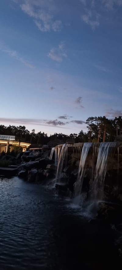 The image shows an outdoor artificial waterfall at dusk, located in the city of Shulgino, Russia. The waterfall cascades over rocks into a pool of water. In the background, there is a building with lights on and a treeline. The sky is a deep blue with some light clouds. The overall scene is serene and tranquil.