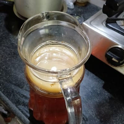 A clear glass pitcher is filled with a brown liquid, topped with a lighter-colored gelatinous mass, possibly a SCOBY for kombucha, sitting on a dark, patterned countertop. Next to the pitcher, on the right, is a stainless steel gas stovetop with a black burner grate and knob. In the background, slightly out of focus, is a silver pot. The location is likely a kitchen, potentially in a residential area. The geotag indicates the location is in Badem, India.