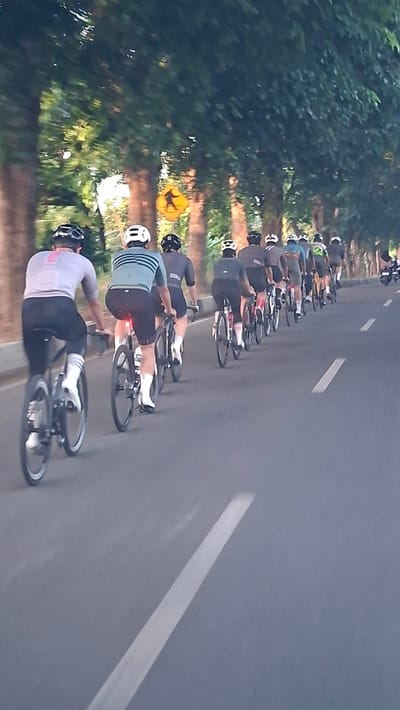 The image depicts a group of cyclists riding on a paved road lined with trees. The cyclists, all wearing helmets and cycling attire, are moving in the same direction down the road. The trees create a canopy effect overhead, with dappled sunlight filtering through the leaves. A yellow pedestrian crossing sign is visible on the left side of the road. The road has white lane markings. The overall scene suggests a group cycling activity in an urban or suburban environment. This scene is located in Santan, Indonesia.