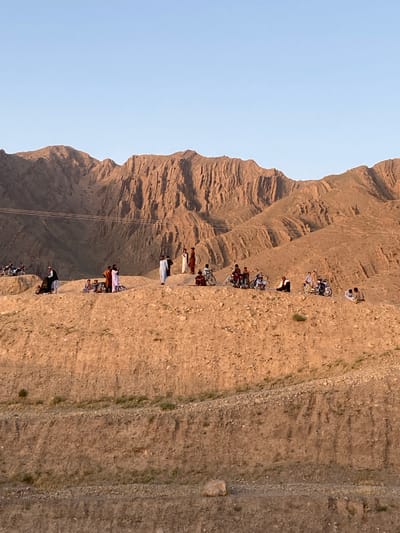 The image depicts a group of people standing and riding bicycles on a narrow dirt path along the side of a rocky, mountainous terrain, likely in a rural setting near Kuchlak, Pakistan. The sky is light blue. The path appears to be cut into the hillside, with steep slopes both above and below it. The people are dressed in various clothing, including traditional Pakistani attire. Some of the individuals are carrying or pushing bicycles along the path. Power lines stretch across the valley from the mountain.
