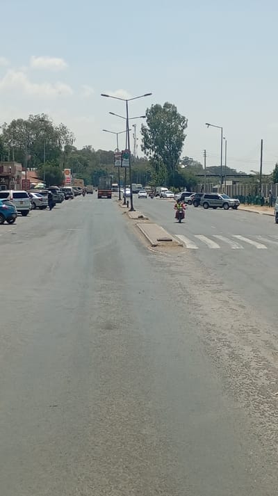 This image displays a daytime street scene in Viwandani ward, Kenya, under a clear sky with scattered white clouds. The primary subject is a multi-lane asphalt road, exhibiting visible wear and tear on its surface.
The road is populated with various vehicles. On the left side, several passenger cars are parked or moving slowly alongside commercial buildings, one of which displays a prominent red and white Shell gas station sign. Further down this lane, a dark-colored truck is observed. On the right side of the road, multiple vehicles, including light-colored SUVs and sedans, are parked.
A pedestrian crosswalk, marked with five white stripes, is visible in the mid-ground on the right side of the road. A person wearing a reflective yellow vest is operating a red motorcycle, positioned directly on this crosswalk in the right lane. Another individual is seen walking on the left side of the road, adjacent to the parked cars.
Multiple streetlights, characterized by their Y-shaped dual arms, are erected along the road, some featuring vertical banners. Raised concrete median strips are present intermittently on the road surface. Dense green foliage from trees and bushes lines both sides of the road and extends into the background. Utility poles with overhead power lines are visible on the right side, and a fence borders the far right edge of the road behind the parked vehicles. The environment suggests an active urban or semi-urban setting.