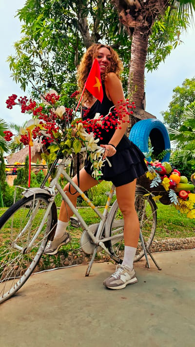 A woman with curly brown hair poses on a white bicycle in Hoa Lư Ward, Vietnam. She is wearing a black dress and white socks with sneakers. The bicycle is decorated with a large bouquet of red berries and white flowers in the front basket and a basket of colorful fruits at the back. A small Vietnamese flag is held up by the woman. The background features lush green trees and foliage, with a clear sky above. A blue tire is visible on the right side of the frame. The scene appears to be outdoors in a tropical or subtropical environment.